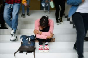 Person sitting on stairs with their head down and arms around knees, appearing distressed while others walk by — representing feelings of anxiety or isolation.